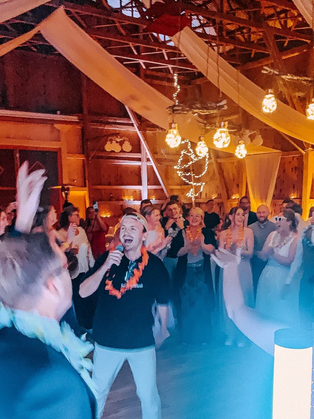 DJ mit Mikrofon animiert jubelnde Gäste mit Blumenketten in einer festlich beleuchteten Scheune